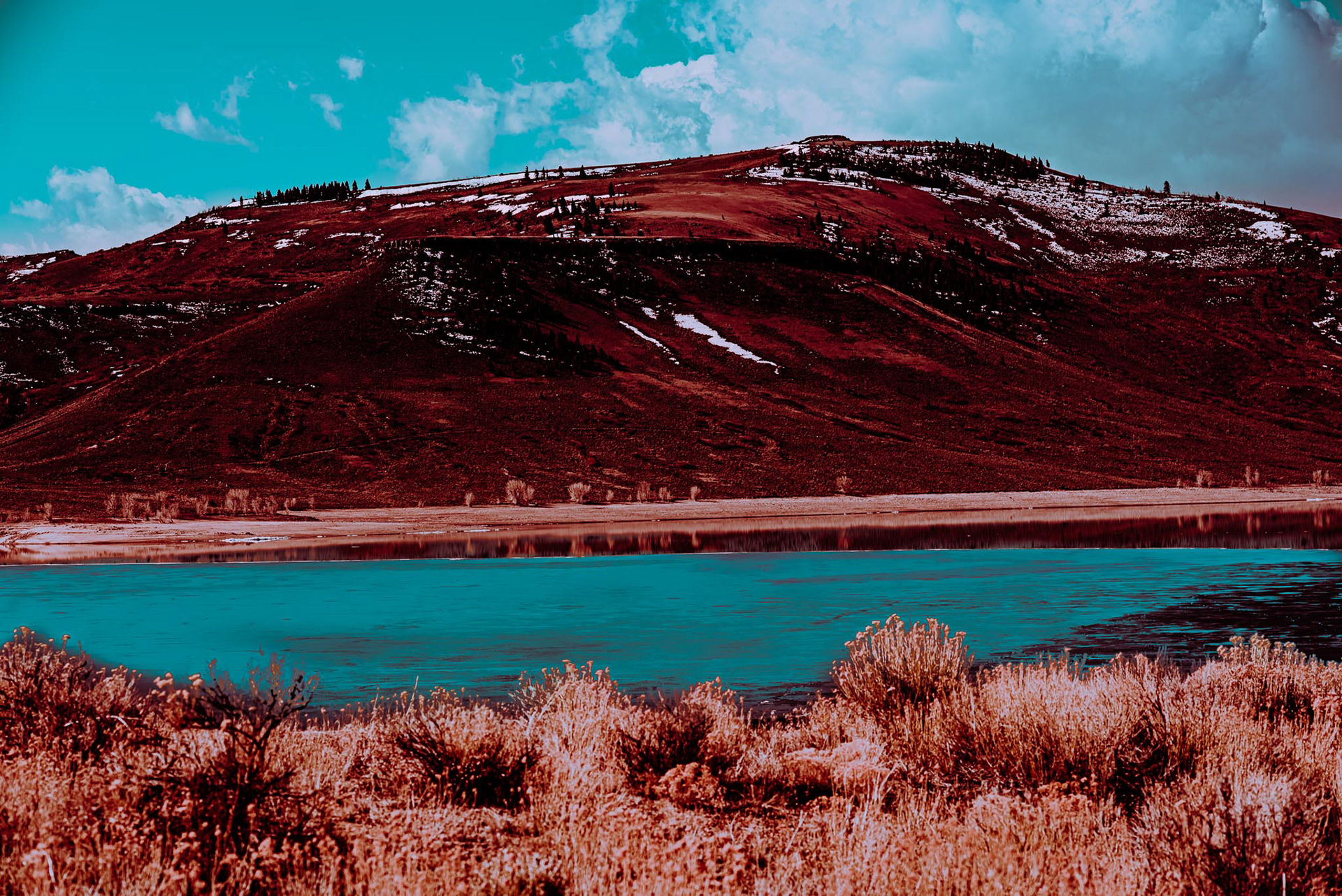 From top to bottom, there is teal skies with white clouds towering over the lightly frosted colorado mountain, overlooking the deep teal water bay below, with warm toned dead weeds.