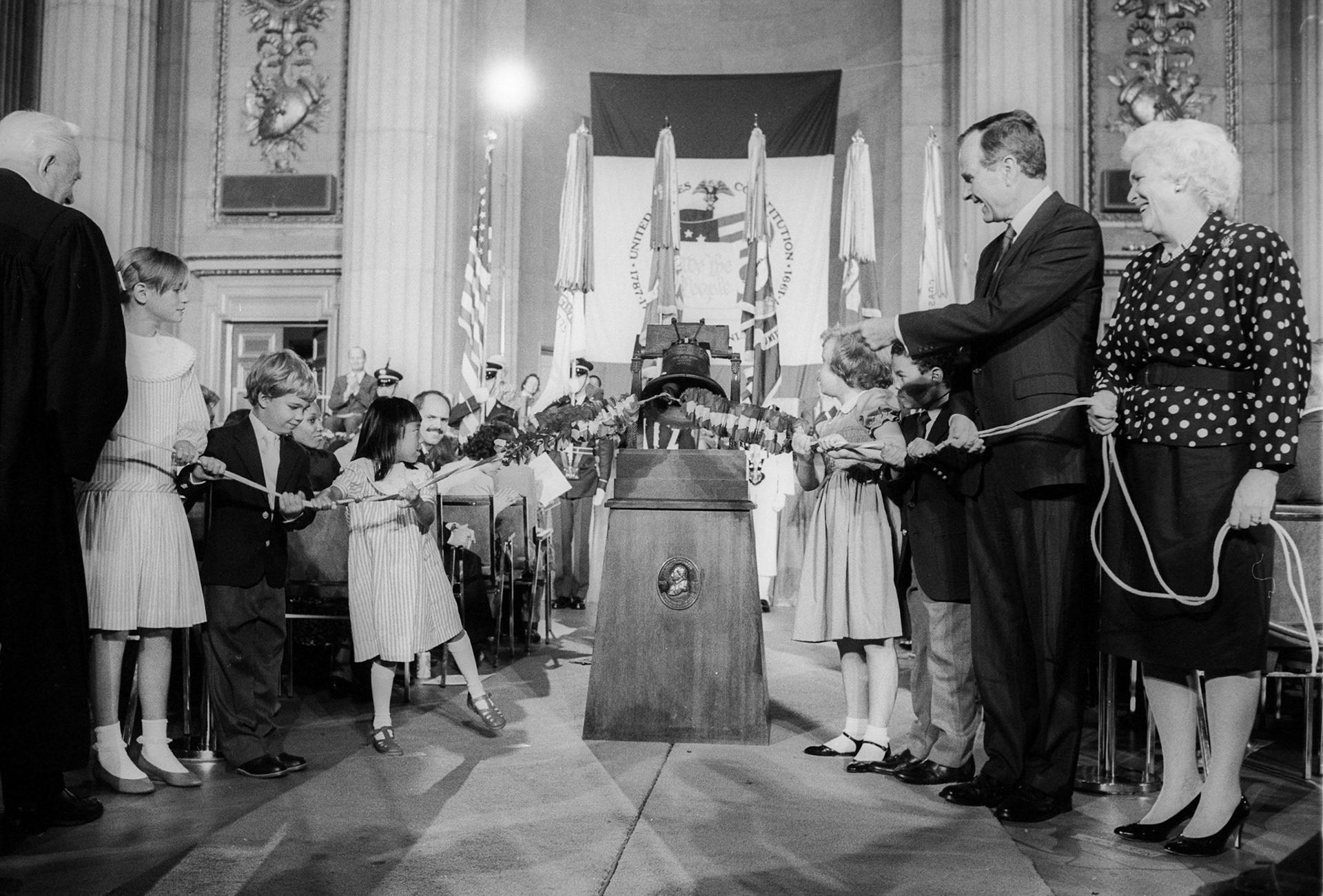 President George H.W. Bush and wife Barbara with school children ring a replica of the Liberty Bell at the swearing-in ceremony of new citizens at the Dept. of Commerce, September 17, 1988.