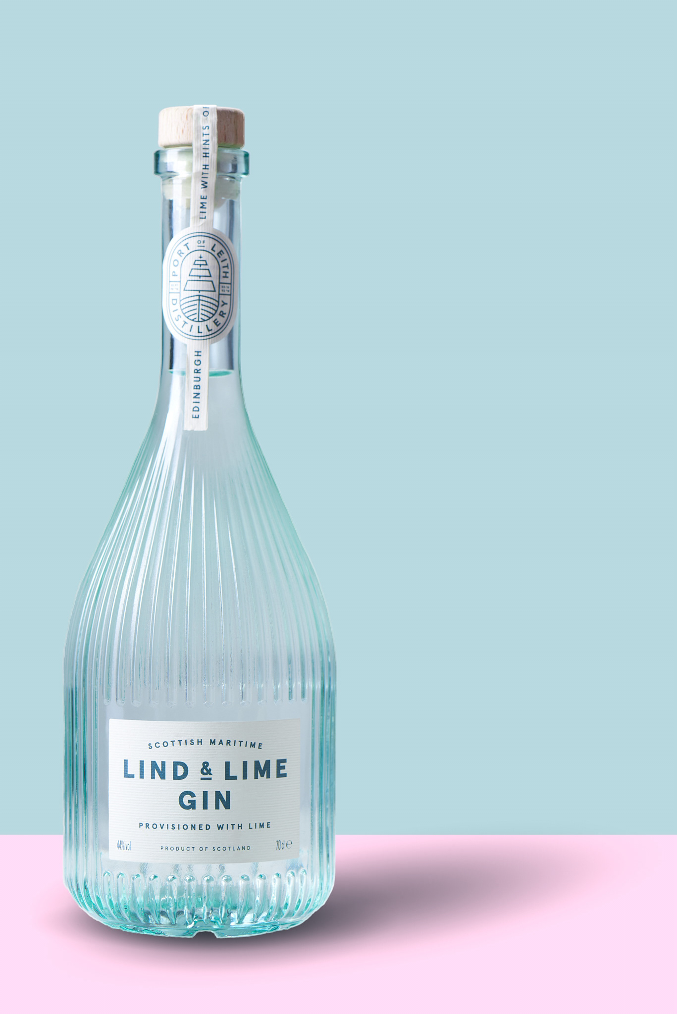 a bottle of Lind and Lime gin, brightly lit, shown against a teal background with a light pink floor, on which the bottle casts a light shadow
