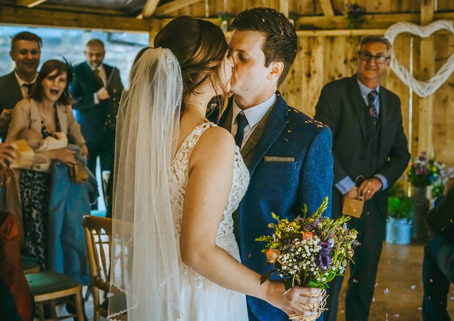 romantic kiss between bride in decorative veiled dress and groom wearing a navy tweed suit at micro barn country wedding surrounded by friends and family