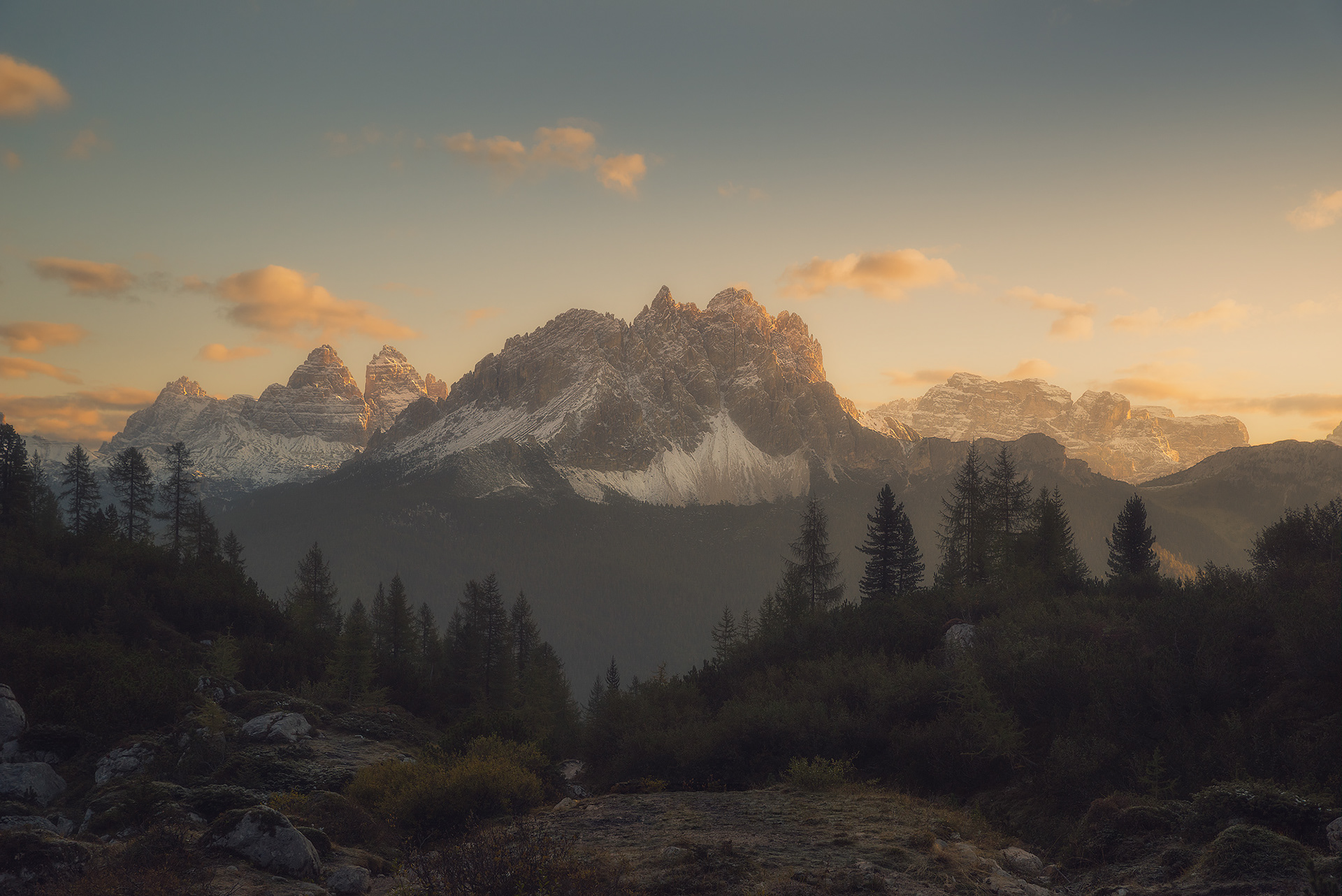 Lever de soleil sur les montages dans les Dolomites en Italie. Des sapins au premier plan puis des pics enneigés teintés d'orange par la lumière du matin. Quelques nuages orangés dans un ciel bleu.